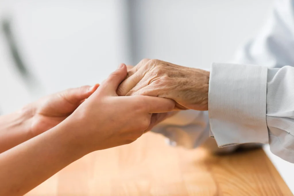 A younger person holds the hands of an older person across a wooden table, both dressed in long-sleeved shirts—sharing comfort and connection as your essential lifeline.