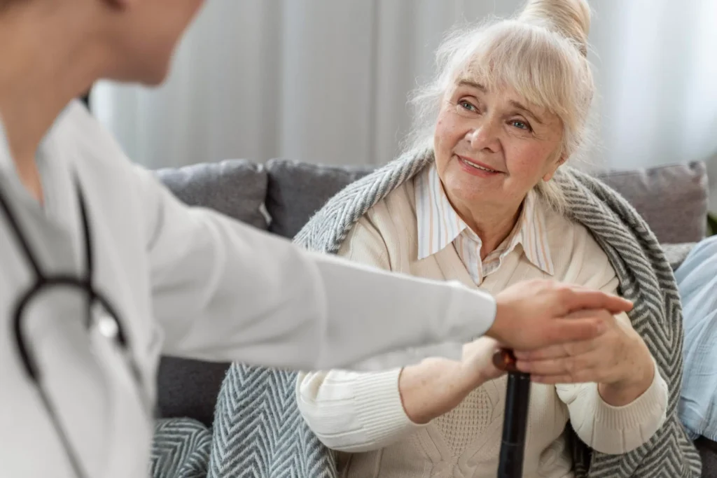 An elderly woman with a blanket over her shoulders smiles while holding a cane, as a healthcare professional in a white coat offers support—highlighting the hidden costs of waiting on Medicaid for essential care.