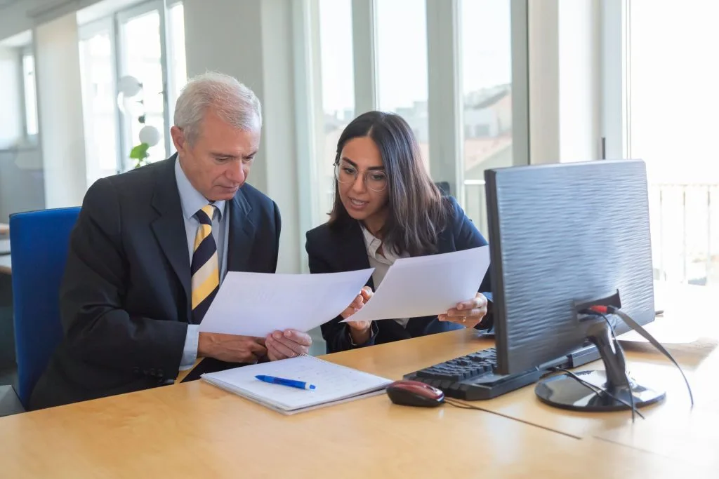 An older man and a younger woman in business attire review documents together at an office desk with a computer, preparing a Maryland Probate Petition.