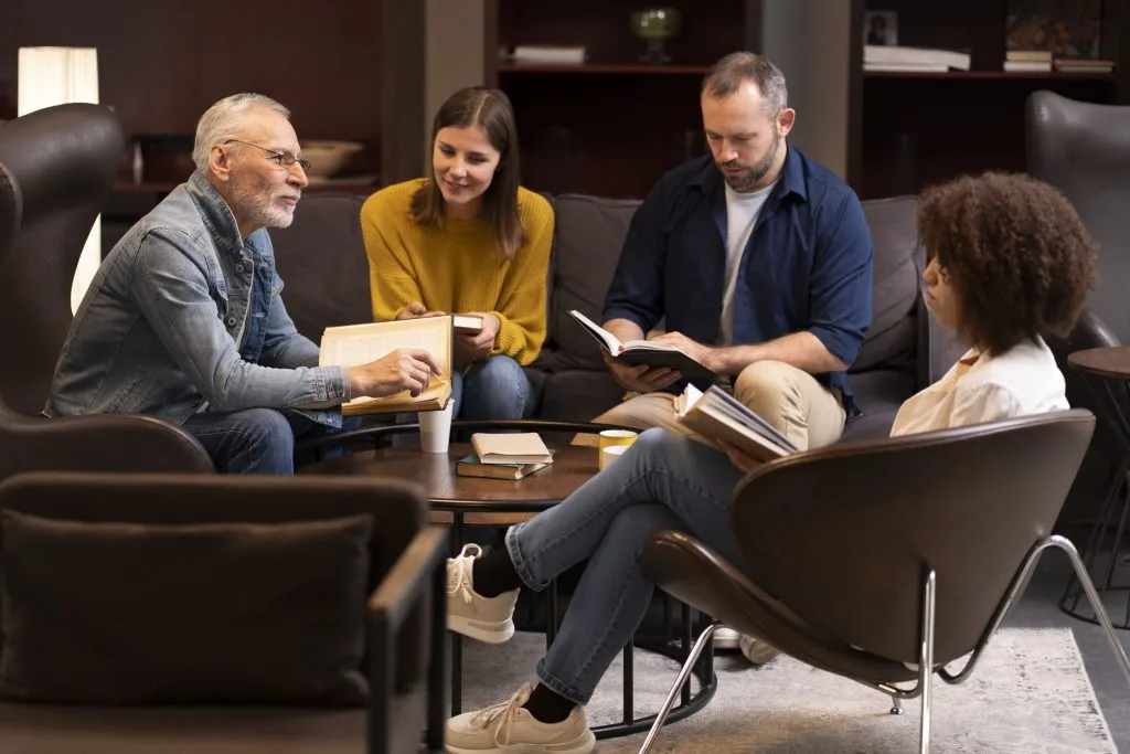 Four people sit in a lounge area, engaged in discussion and reading books around a coffee table with notebooks and coffee cups, sharing ideas on how to minimize family conflicts after your passing.