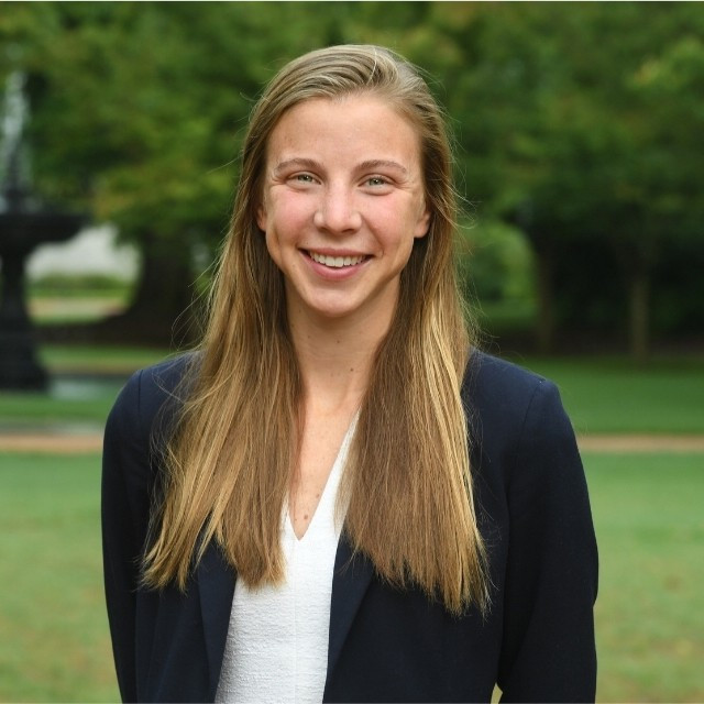 Lydia, hosting Sawyer & Associates, stands outdoors with long blonde hair in a navy blazer and white top, smiling warmly amid greenery and a blurred fountain in the background.
