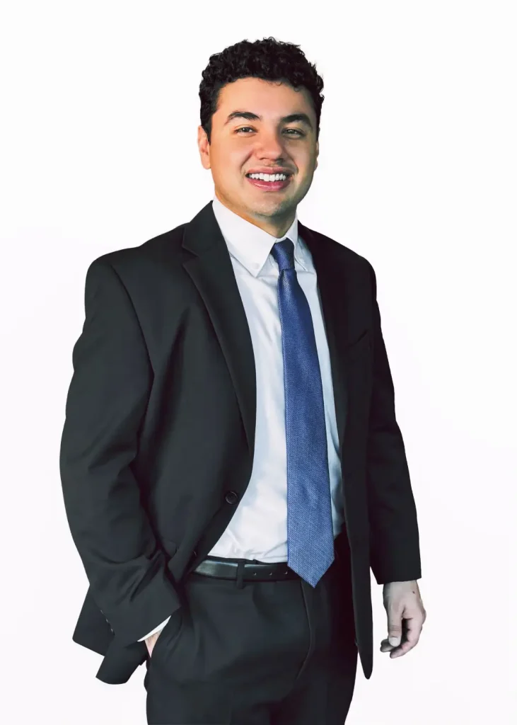 Joe Bendekovic of Sawyer & Associates stands against a plain white background, smiling in a black suit, white shirt, and blue tie with one hand in his pocket.