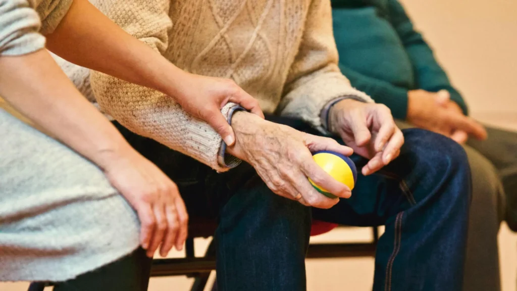 A person in a sweater holds a small yellow and blue ball while another gently places a hand on their forearm, offering comfort during a medicaid crisis planning discussion; both are seated.