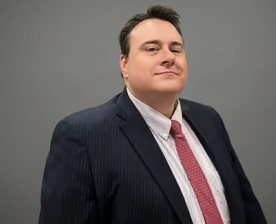 A man from Nick Wilson Sawyer & Associates, dressed in a dark pinstripe suit, white shirt, and red patterned tie, stands confidently before a plain gray background, looking directly at the camera.