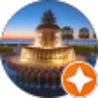 A lit fountain with water cascading at sunset, with palm trees and a body of water in the background; an orange star badge is in the lower right corner.