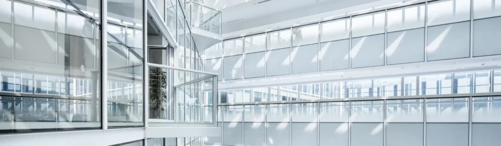 Interior view of a modern building with large glass windows, white walls, and sunlight casting diagonal shadows across surfaces.