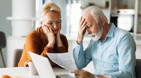Two older adults sitting at a table look concerned while reviewing a document in front of a laptop.