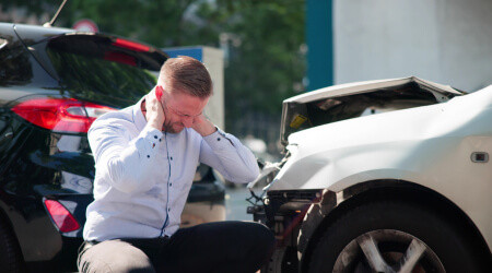 A man sits on the ground with his head in his hands beside two cars with damaged front ends, indicating a recent collision.