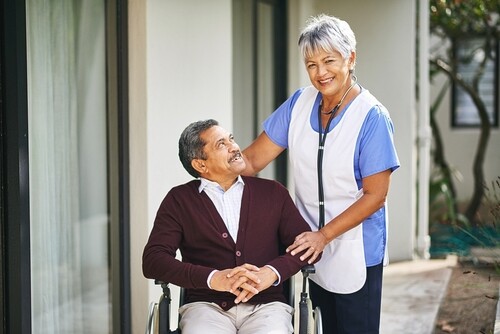 senior man in wheelchair smiling with caregiver wearing stethoscope outdoors